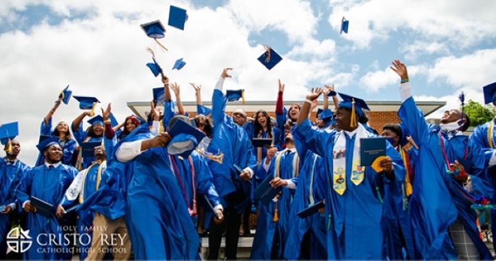 A group of graduating students from Holy Family Cristo Rey Catholic High School in blue gowns joyfully throwing their caps into the air.