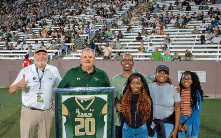 A group of six smiling people posing on a football field in front of a stadium crowd, with one man holding a framed green UAB football jersey.