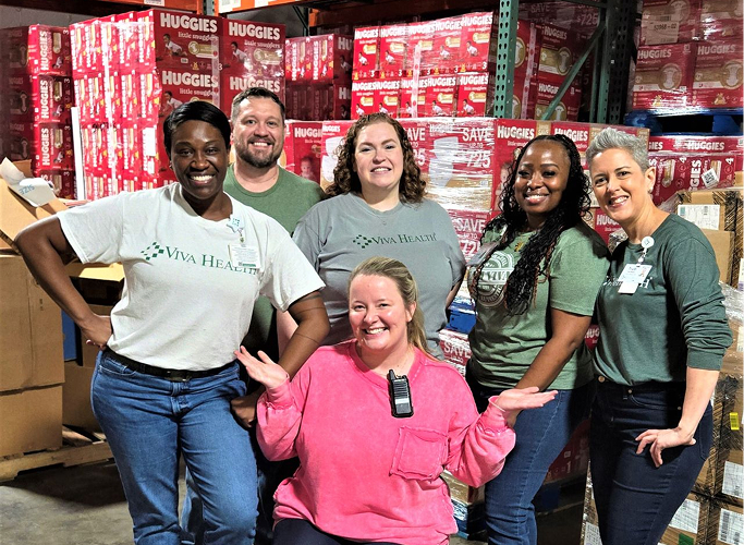 A group of volunteers posing in front of tall stacks of diaper boxes in a warehouse.