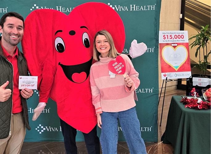 Two people posing with a large red heart mascot at a Viva Health event.