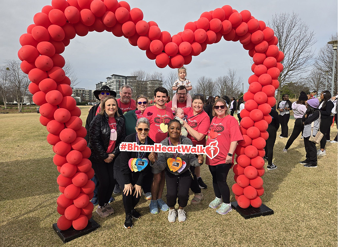A group posing inside a large red balloon heart archway holding a Birmingham Heart Walk sign.