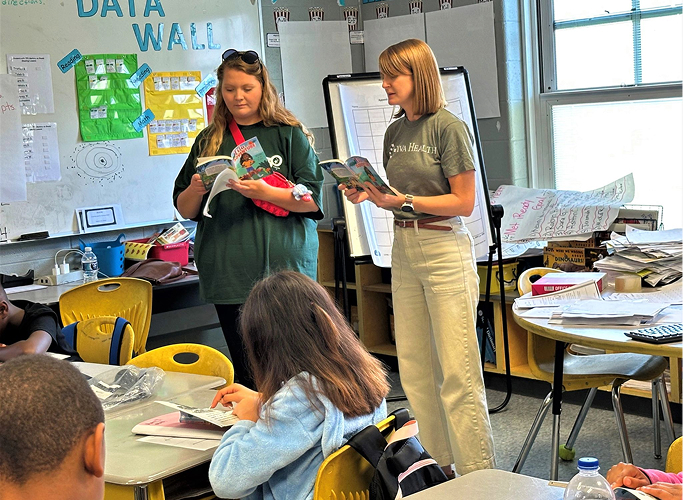 Two volunteers standing in a classroom reading books to young students.