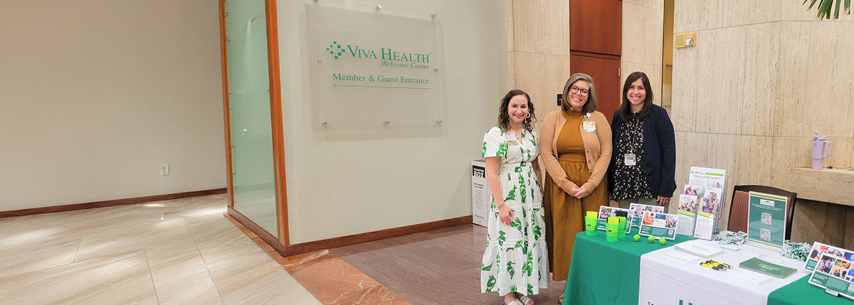 Three smiling women standing next to a promotional table in front of a glass Viva Health Welcome Center sign.